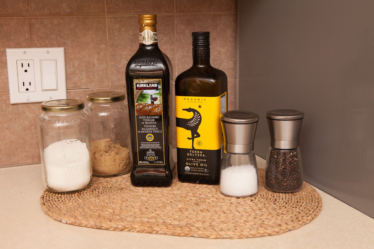 A kitchen countertop is neatly arranged with various cooking essentials. Two bottles of olive oil are positioned prominently alongside jars filled with flour and brown sugar. Salt and pepper shakers rest on a woven placemat, creating a functional yet appealing kitchen space.