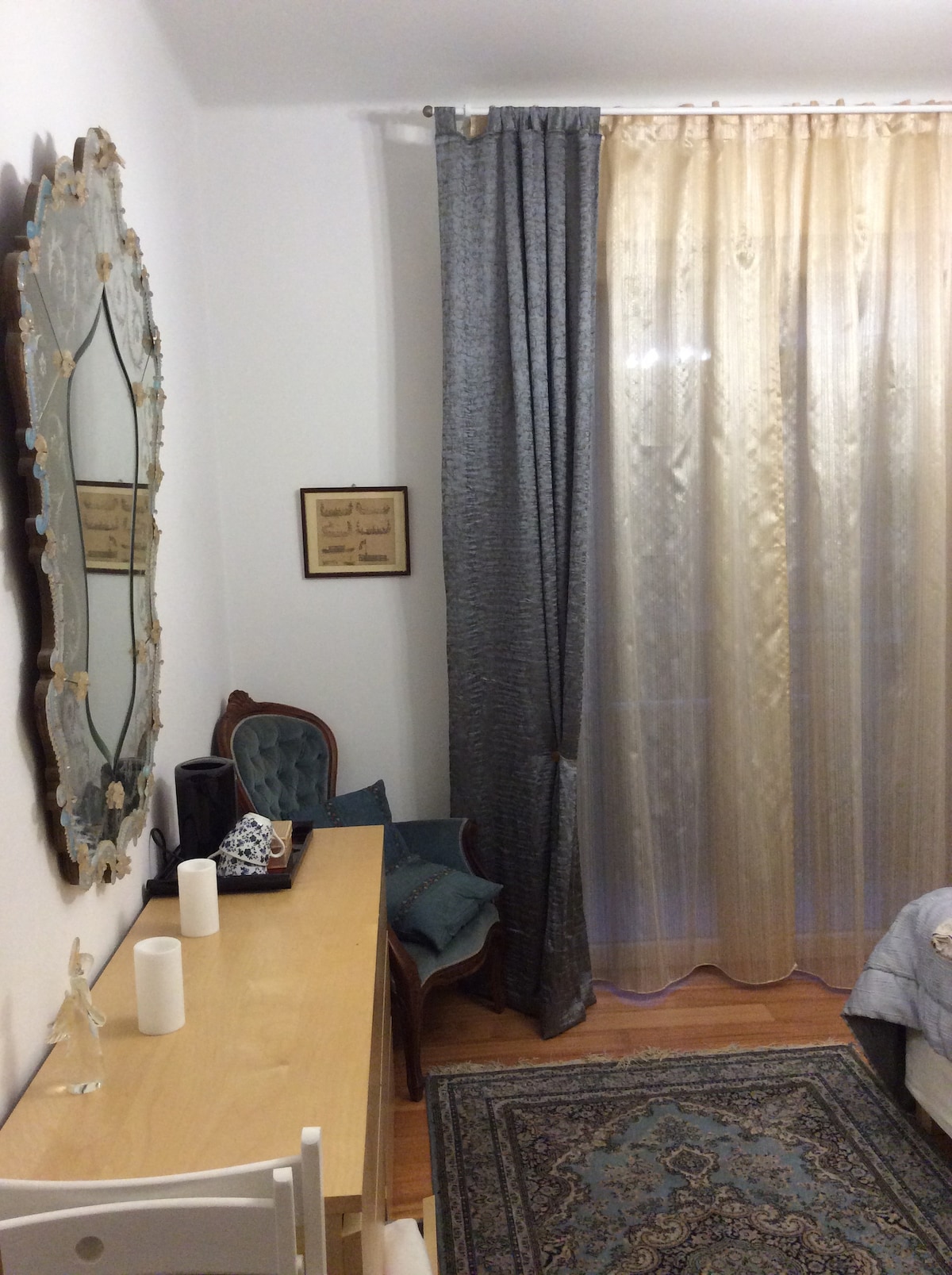 A cozy room features a wooden desk accompanied by an elegant chair with a vintage design. A large mirror with ornate detailing reflects natural light from sheer curtains. A decorative rug adds warmth, while candles provide a subtle decorative touch.
