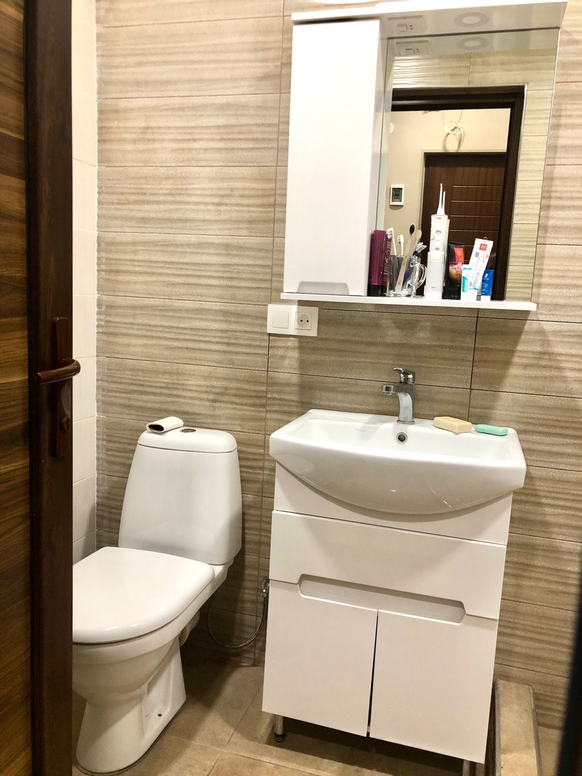 A compact bathroom is shown, featuring a white toilet and a sleek sink with an under-sink storage cabinet. Above the sink, a mirrored cabinet is visible, showcasing personal care items. Neutral-toned walls add to the clean, modern aesthetic.