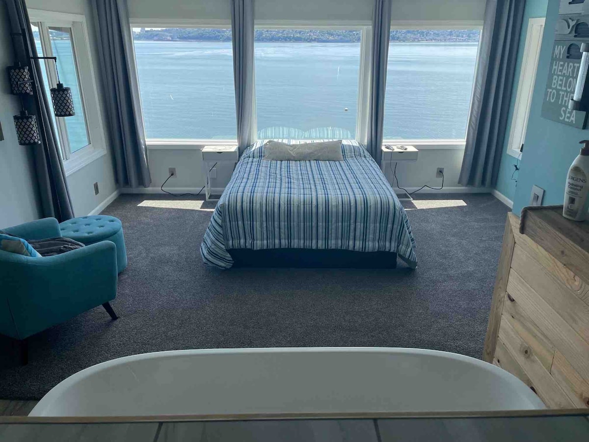 A bright bedroom features a spacious bed dressed in a striped blue and white blanket, positioned near large windows showcasing sweeping views of the water. A cozy chair in a soothing blue tone complements the calming decor, while natural light fills the space.