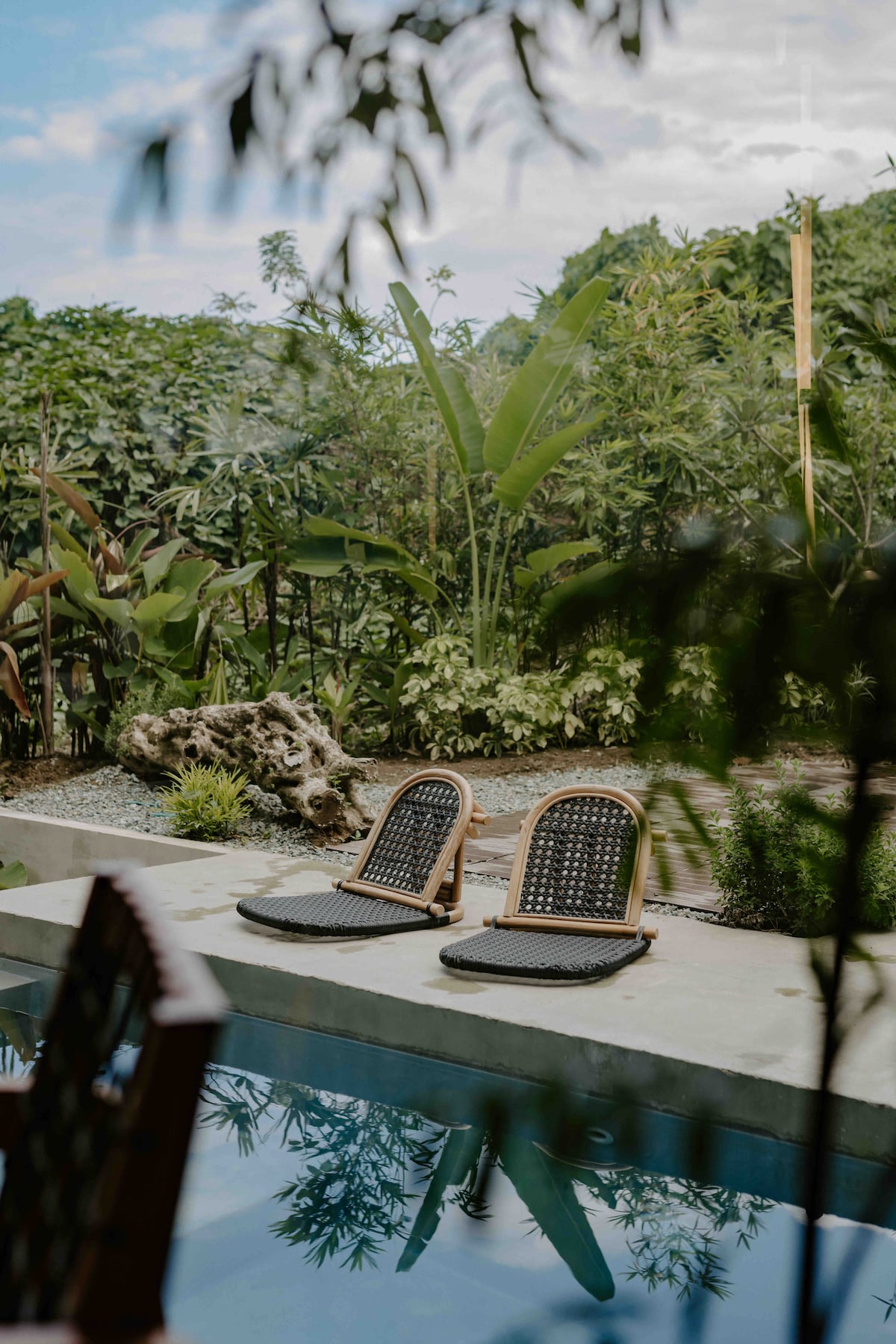 Two lounge chairs with woven seats are positioned near a serene infinity pool, reflecting the surrounding lush greenery. Tall tropical plants provide privacy, while a natural wooden feature is visible in the landscape, adding to the tranquil atmosphere.