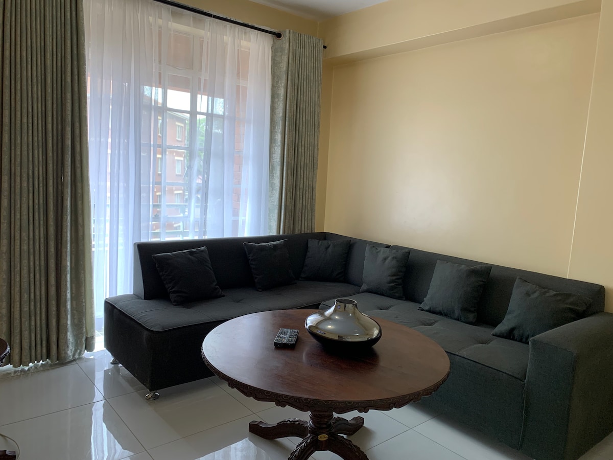 A spacious sitting area is presented with a dark sofa arranged around a round wooden table. Sheer curtains allow natural light to filter in, enhancing the warmth of the interior. A remote control is placed atop the table, providing convenience for entertainment options.