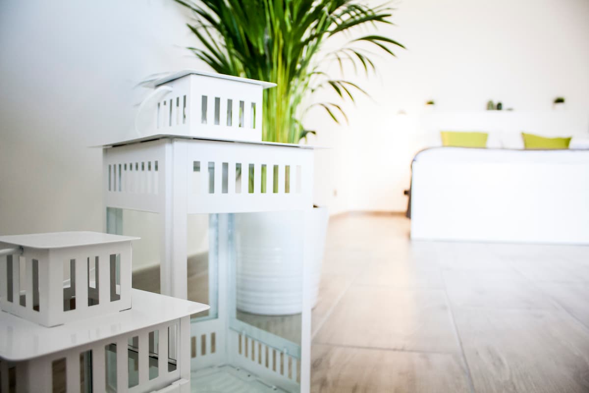 A clean and modern space is presented, featuring white decorative lanterns of varying sizes positioned near a large green plant. In the background, a spacious bed with green accents is visible, emphasizing a calming and airy atmosphere.