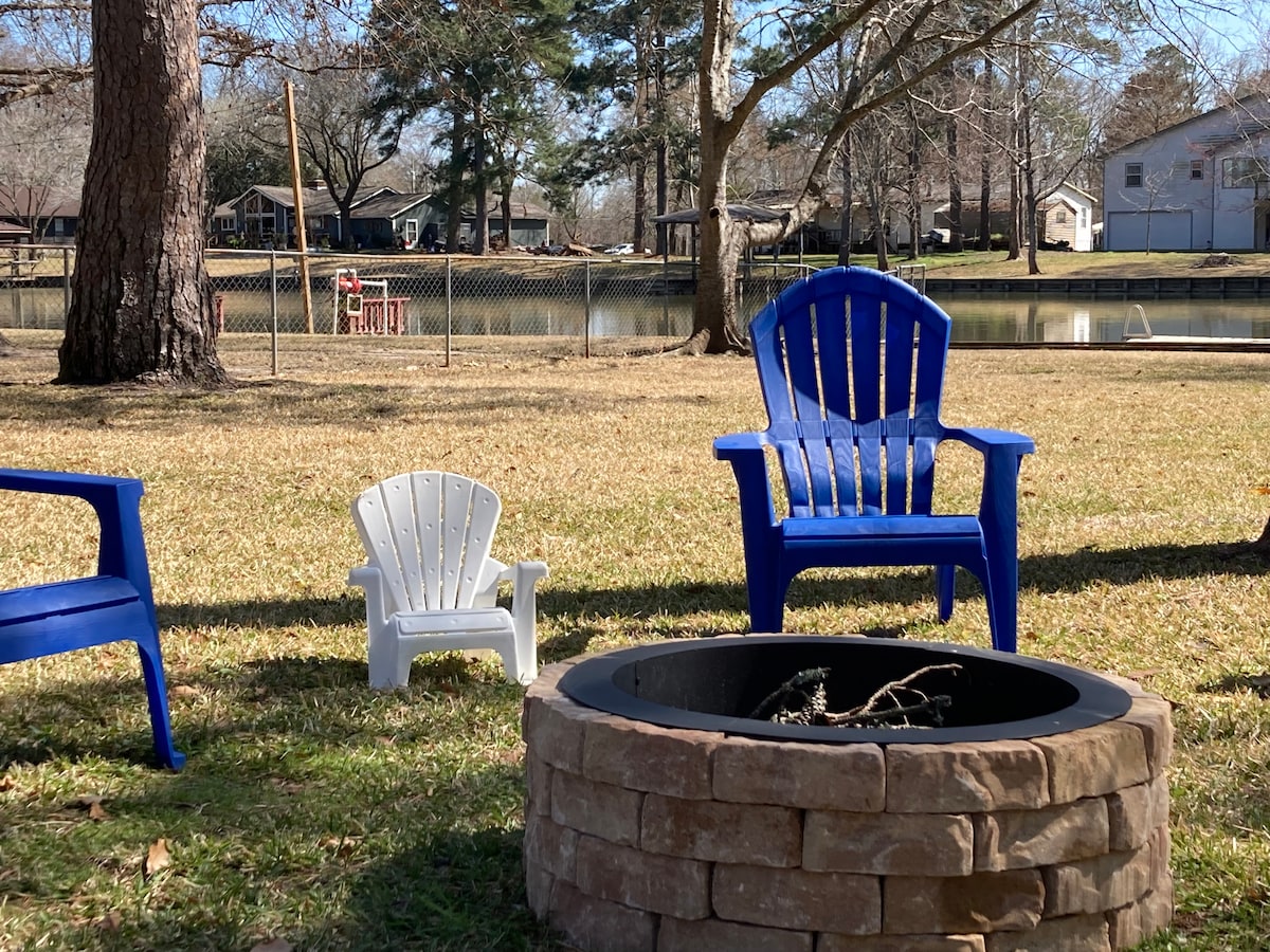 A fire pit constructed of stone is surrounded by blue lawn chairs and a small white chair. The grassy area leads to a tranquil view of the lake, framed by trees and a fence in the background, creating a relaxed outdoor setting.