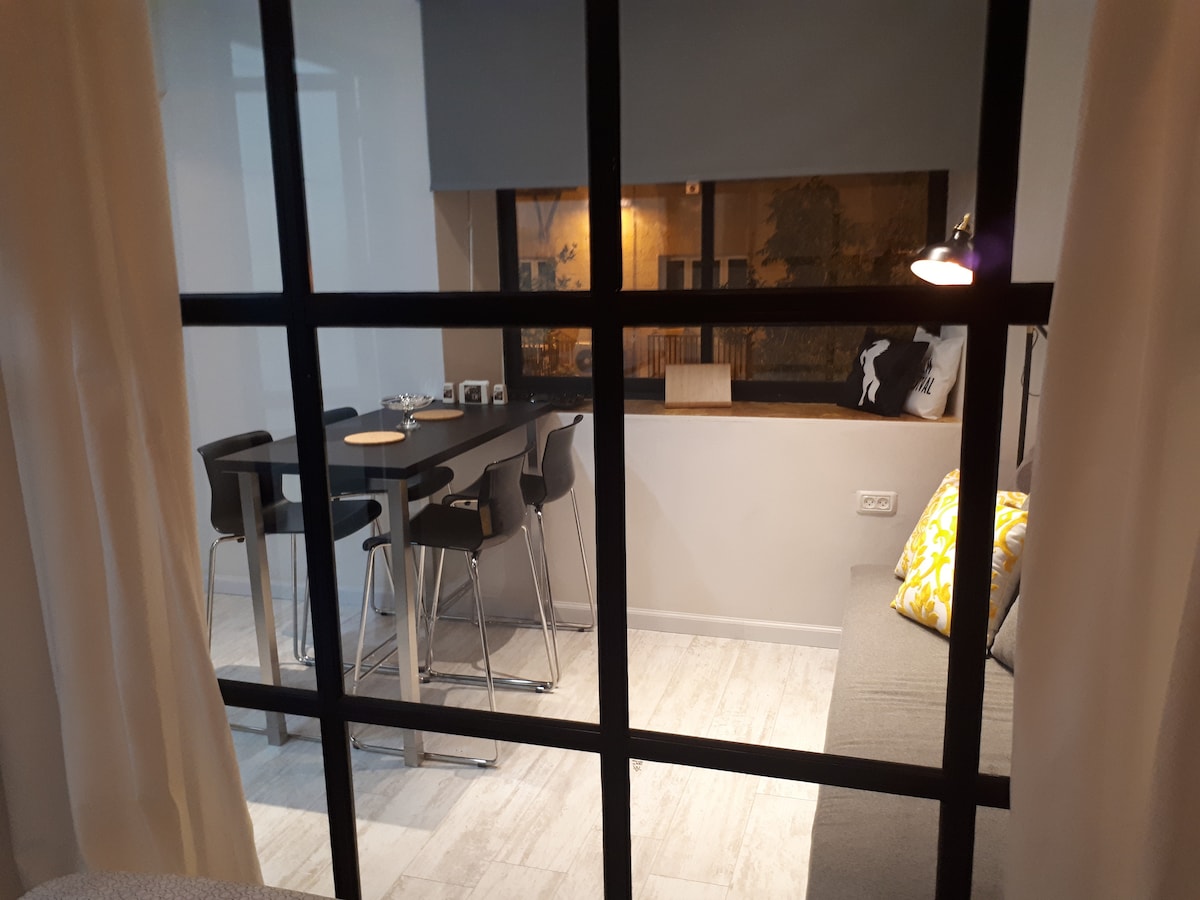 A modern dining area is visible through a large window, featuring a sleek table surrounded by four black stools. A gray sofa is positioned nearby, adorned with patterned cushions. Soft lighting from a wall-mounted lamp creates a comfortable ambiance.