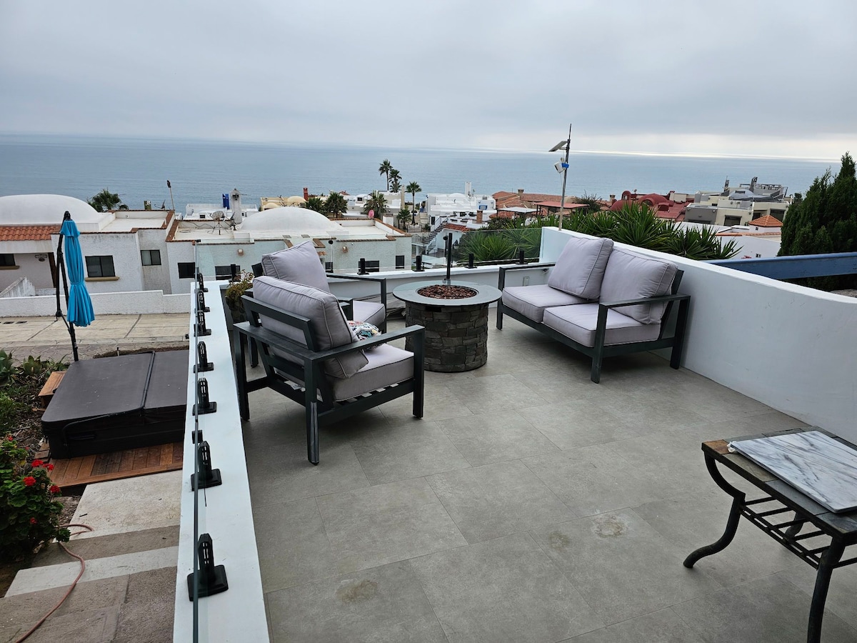 A spacious outdoor seating area features two comfortable chairs and a small fire pit, surrounded by a stone border. A table sits nearby, with an umbrella providing shade. Views of the ocean and the neighboring rooftops create a calming atmosphere.