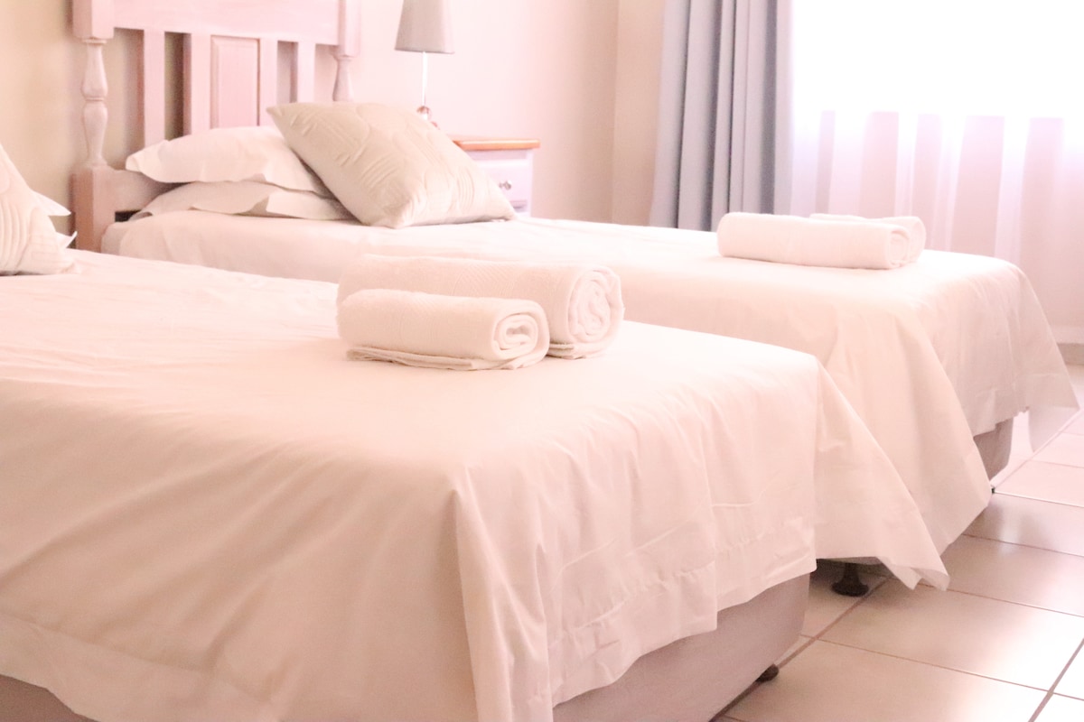The room features two spacious beds, each neatly made with white linens. Rolled towels are placed at the foot of each bed. Soft natural light enters through a large window draped with light curtains, illuminating the light-colored decor.