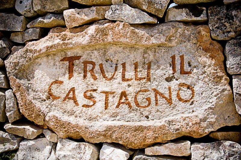 A rustic stone sign engraved with the words 'Trulli Il Castagno' is set against a backdrop of weathered stones. The natural texture and earthy tones of the sign reflect the traditional architecture of the surrounding trulli.
