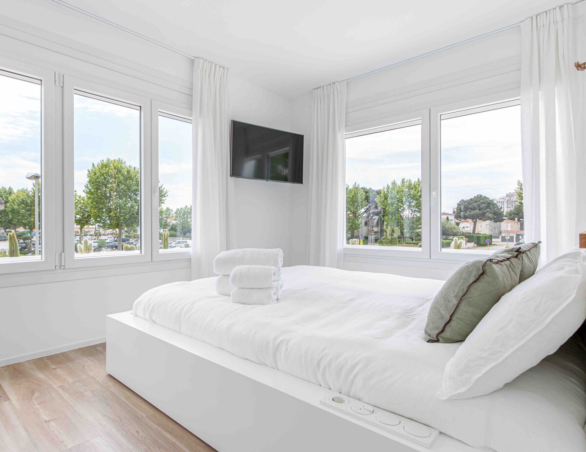 A bright bedroom features a spacious bed with crisp white linens and two neatly rolled towels placed on it. Large windows with sheer curtains offer views of the greenery outside, and a flat-screen TV is mounted on the wall for additional comfort.