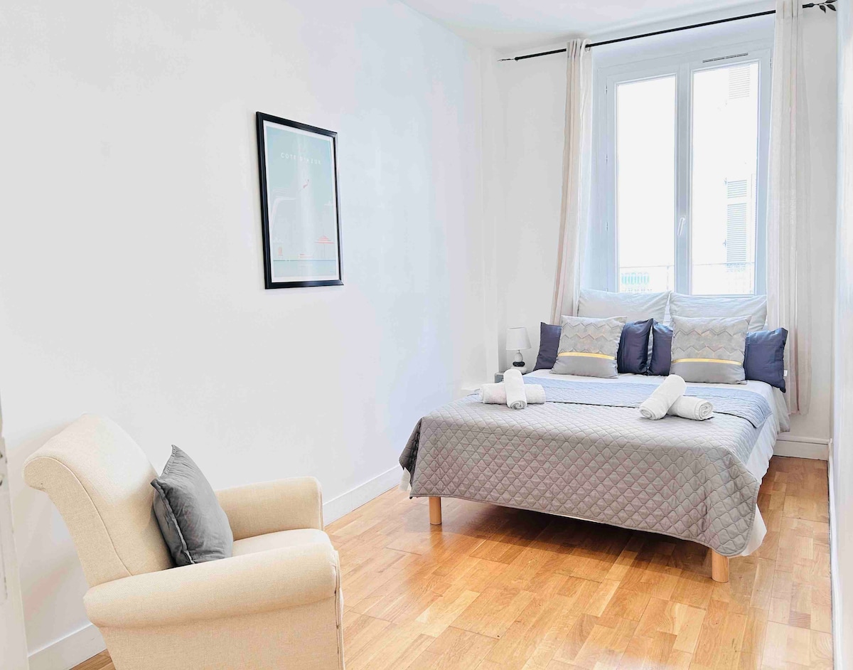 A bright bedroom features a spacious bed with neatly arranged towels. A comfortable armchair sits nearby, while large windows allow natural light to fill the room. Light-colored walls and wooden flooring contribute to a clean and inviting ambiance.
