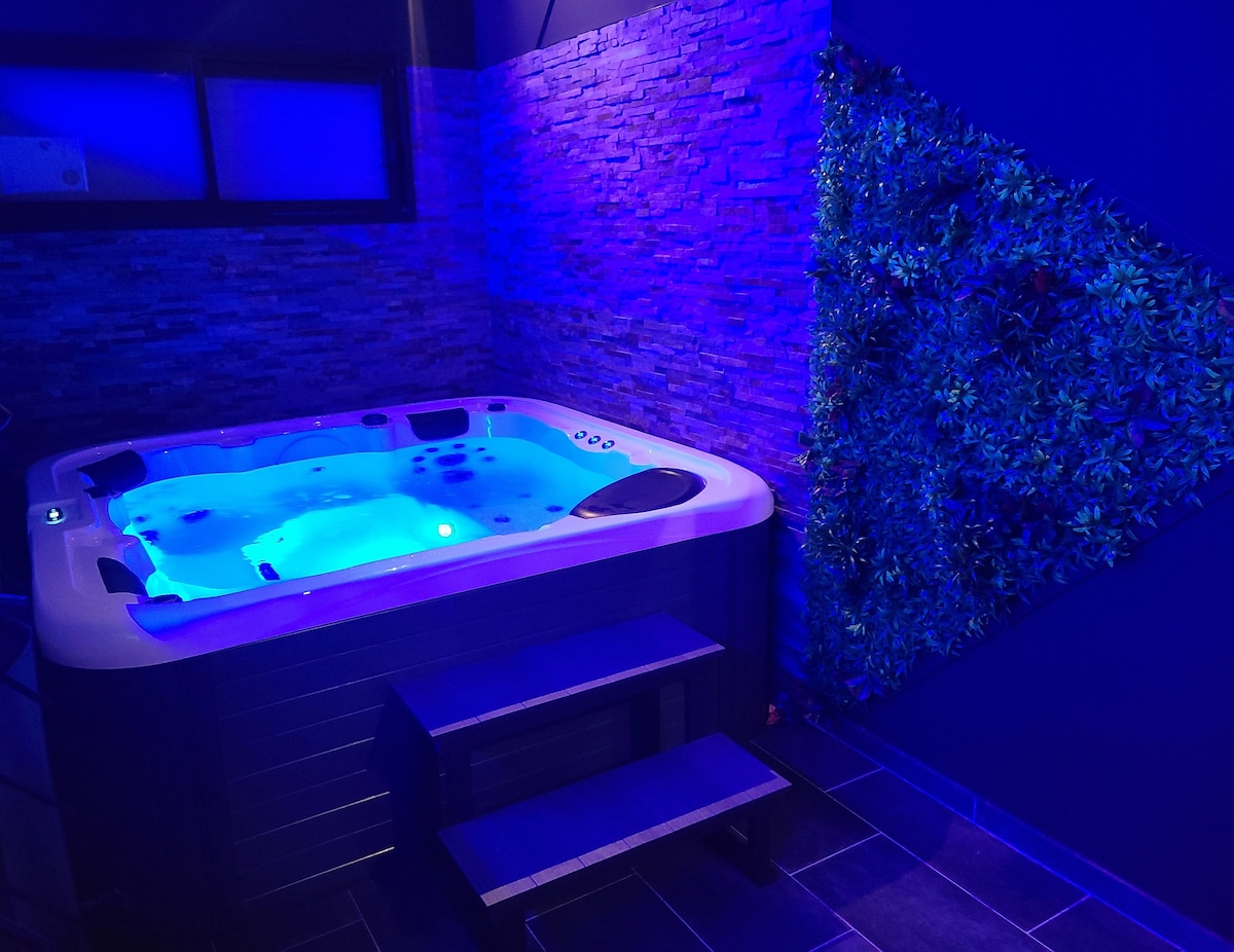 An indoor jacuzzi is set against a backdrop of textured stone walls illuminated in soft blue lighting. Greenery is integrated into the design, adding a touch of nature. Steps lead up to the jacuzzi, creating a relaxing and inviting atmosphere.
