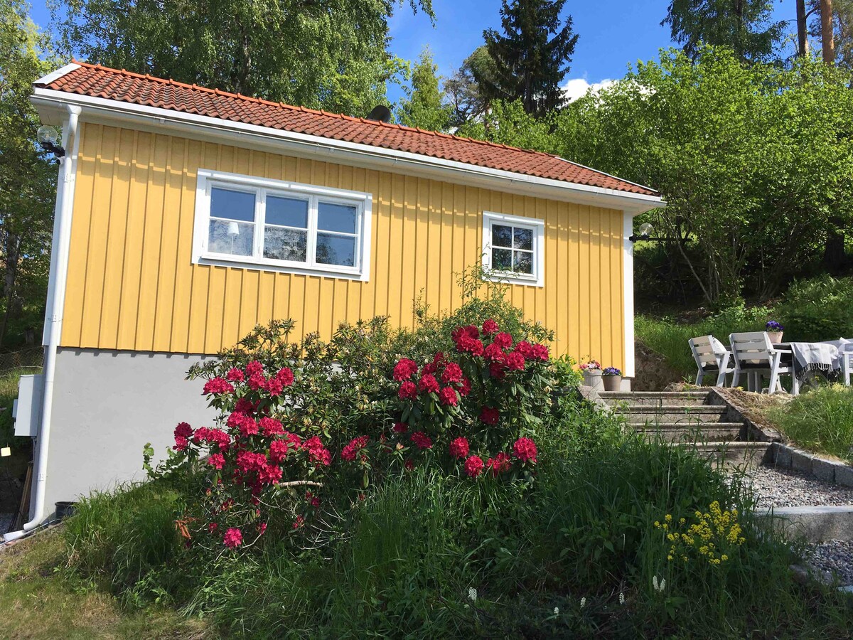 A charming yellow house is set against a backdrop of lush greenery. The lower part features a gray foundation, while blooming flowers add color in front. A stone pathway leads to a patio equipped with light-colored outdoor furniture, inviting relaxation in a tranquil setting.