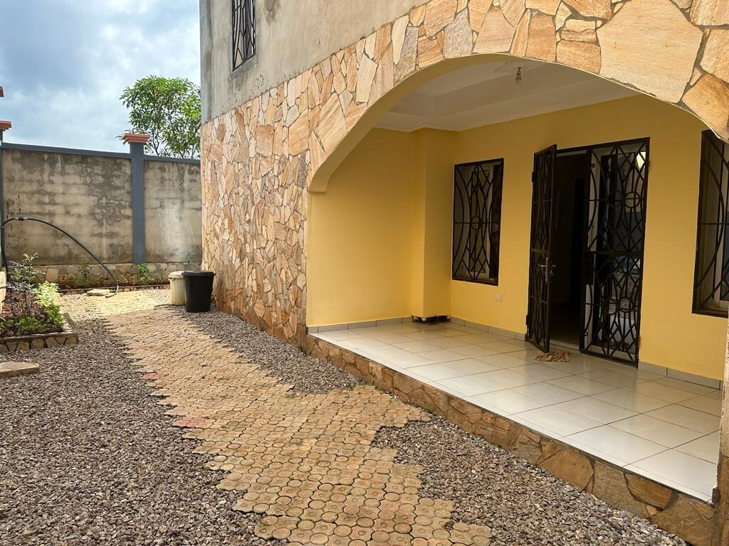 An outdoor area features a stone pathway leading to an arched entrance. The wall is adorned with a combination of large stones and smooth surfaces, while green plants are placed along the borders. Light gray tiles extend across the entrance, complemented by black wrought iron doors.