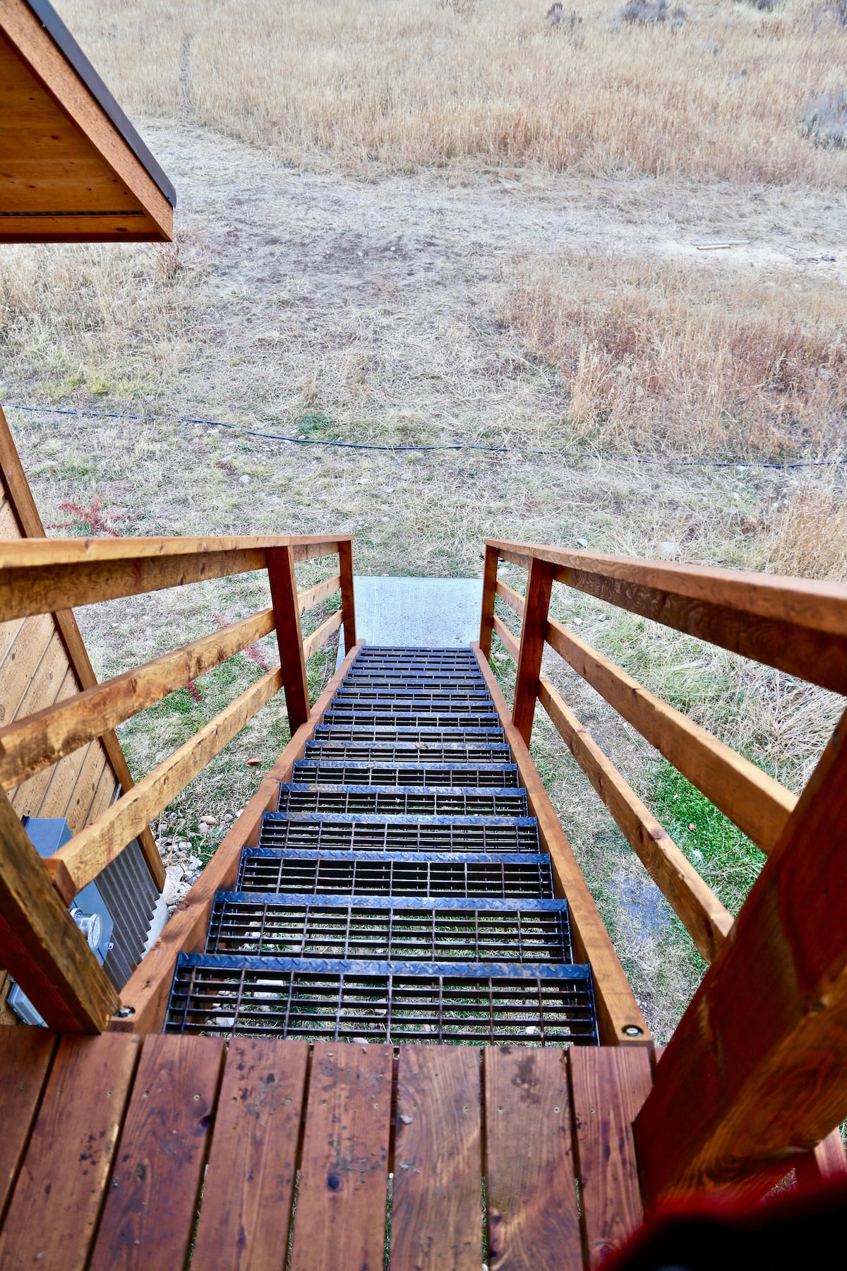 A metal grate staircase leads down from a wooden deck, surrounded by railings. Below, a grassy landscape stretches out, with dried grass visible in the foreground. The scenery beyond is open and natural, inviting exploration.