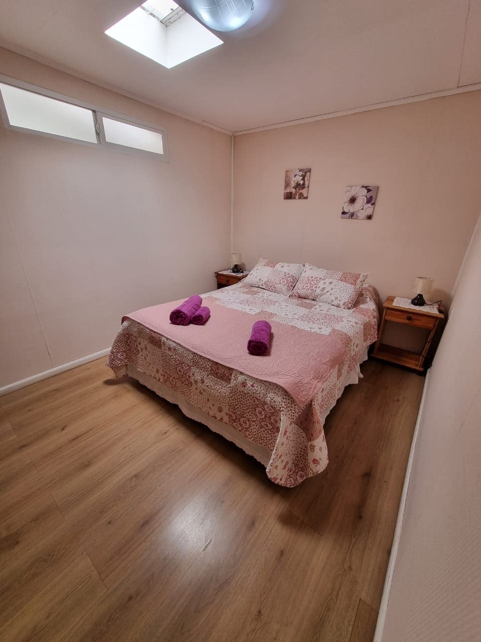 A bright sleeping space features a spacious bed covered with a patterned blanket and decorative pillows. Two towels are neatly placed at the foot of the bed. Natural light enters through a small window and a skylight above, enhancing the warmth of the room.