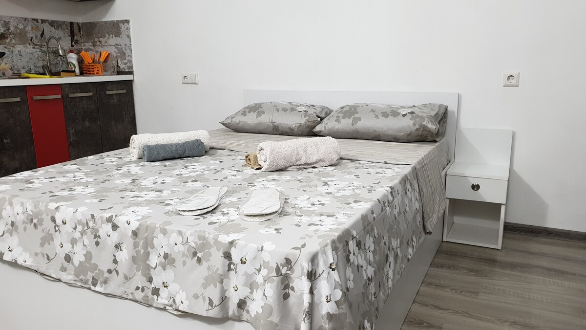 A comfortable sleeping area features a spacious bed with floral-patterned bedding and two pillows. Fresh towels are neatly arranged on the bed, alongside a small nightstand. The kitchen area is visible in the background, with cabinetry and countertop appliances.
