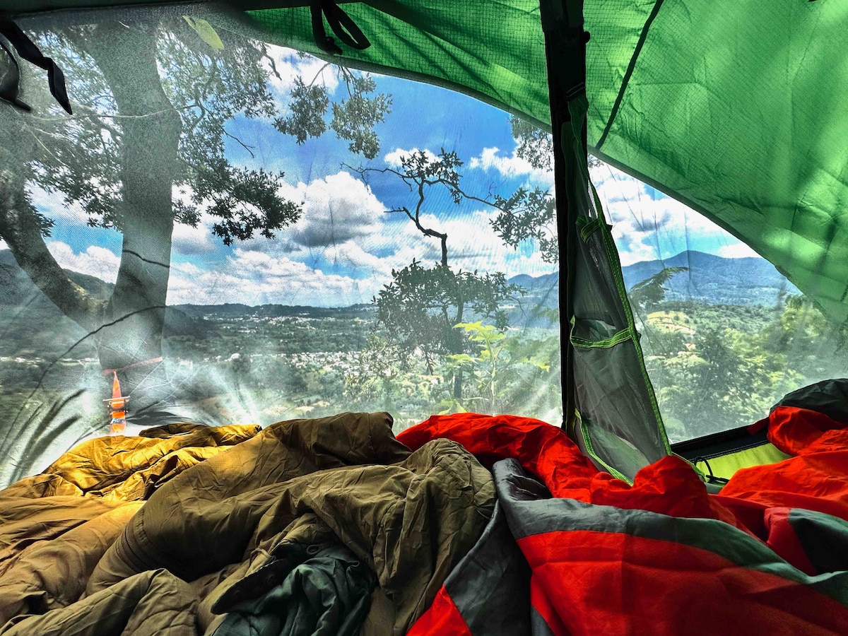 An interior view of a suspended tent showcases colorful sleeping bags laid out on the bedding. The mesh panels offer an unobstructed view of the surrounding landscape, featuring green trees and distant mountains under a partly cloudy sky.