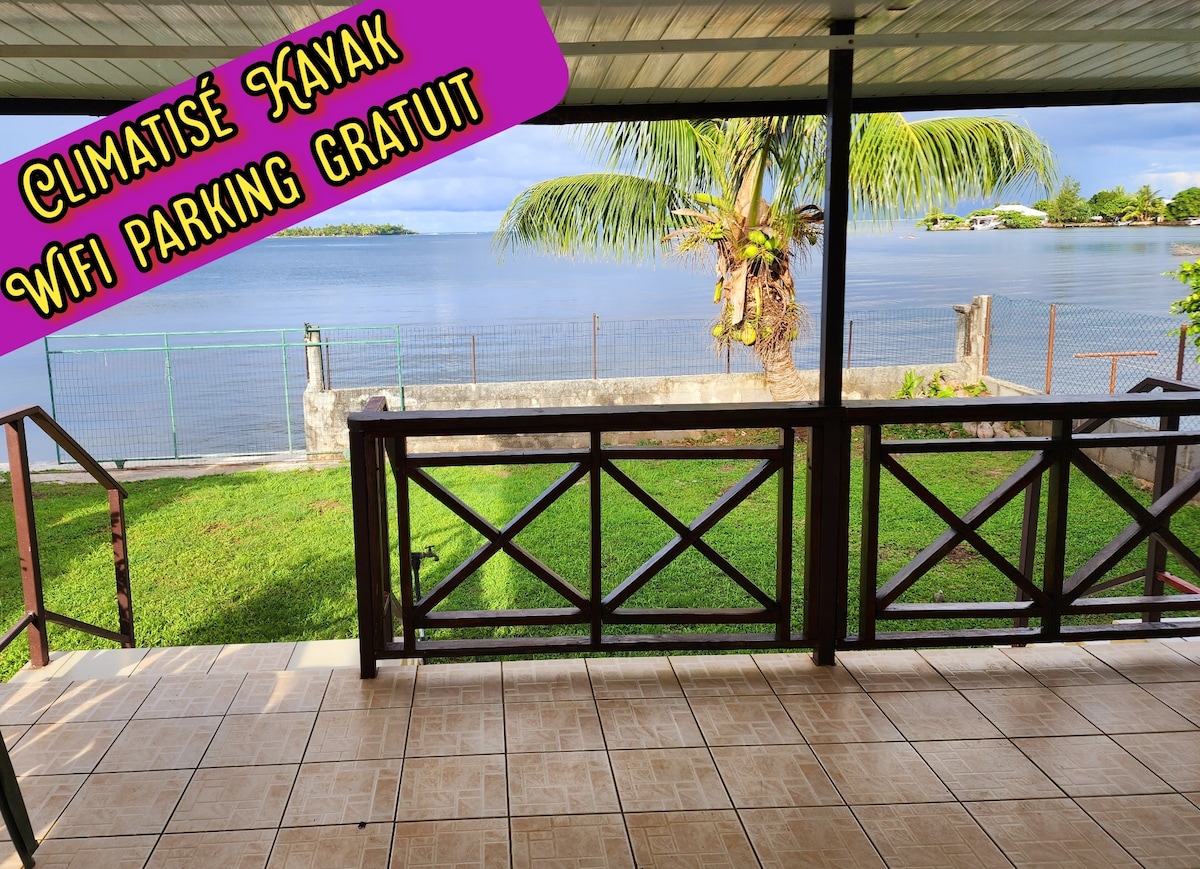 A covered terrace features tiled flooring and a view of the waterfront, with a green lawn leading to the water's edge. A palm tree adds a tropical touch, and the general area suggests a relaxing outdoor space for guests.