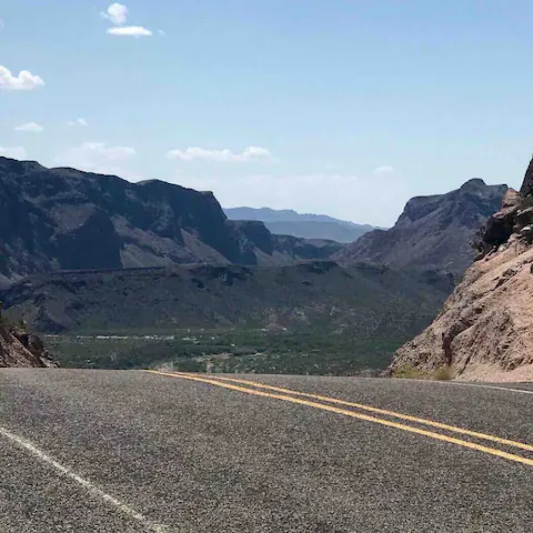 A winding road stretches through a rugged landscape, flanked by towering rocky mountains. The expansive valley below is visible, showcasing the diverse terrain and distant mountain ranges under a bright blue sky. Soft, fluffy clouds dot the horizon.