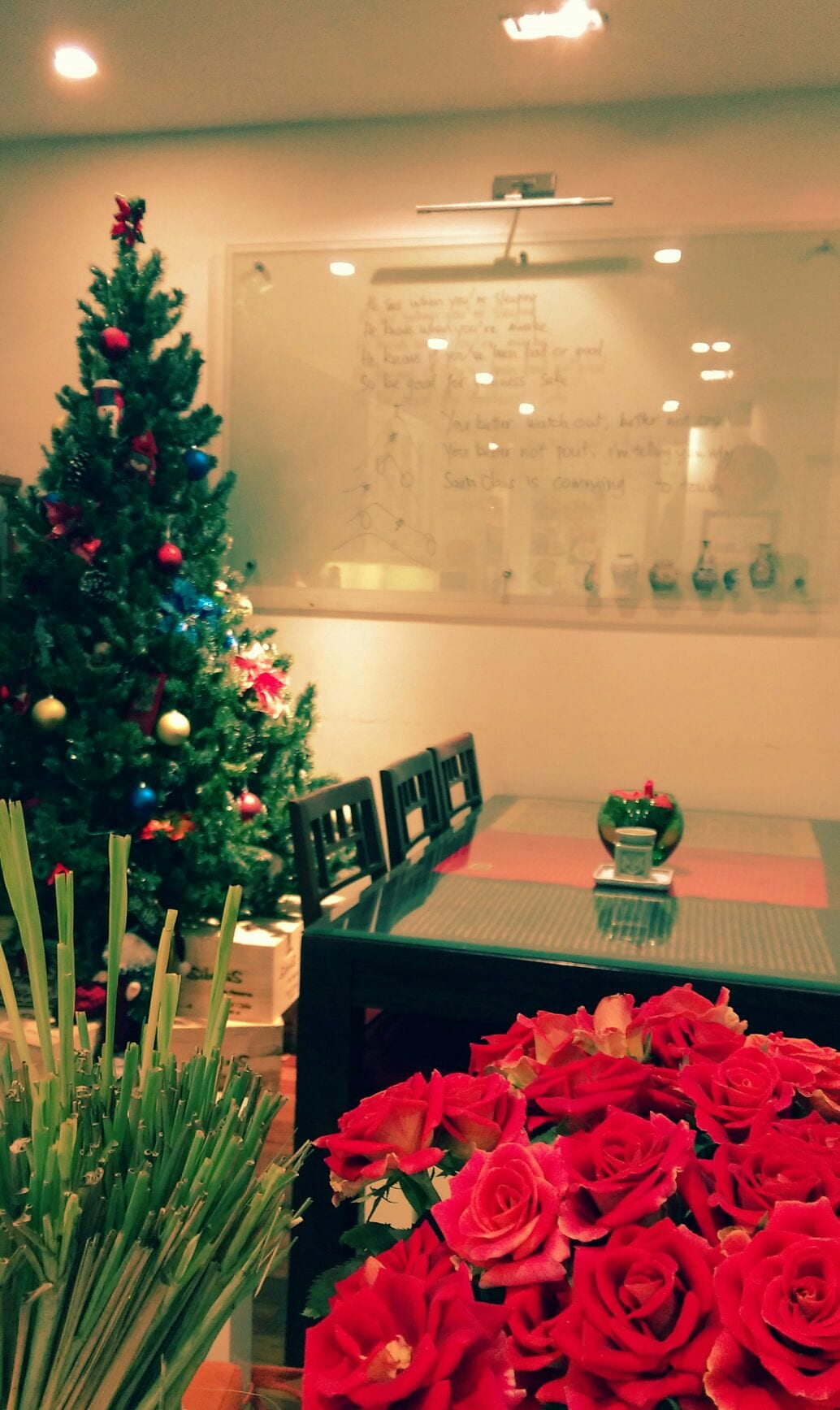 A dining area is presented with a dark wood table surrounded by chairs. A Christmas tree, adorned with ornaments, adds a festive touch. A bouquet of red roses is positioned in the foreground, complemented by a stylish centerpiece on the table. A glass whiteboard is mounted on the wall.