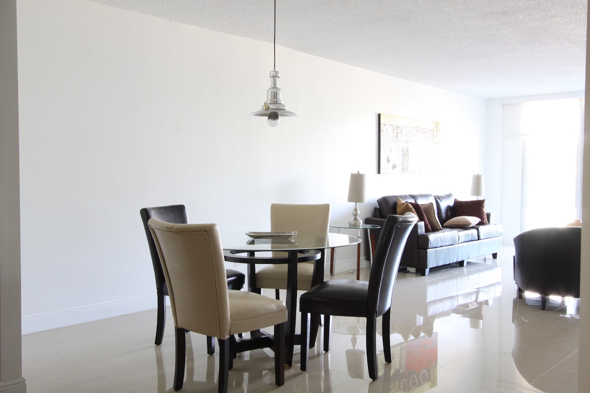 A dining area is set with a circular glass table surrounded by four chairs, two in light tones and two in dark tones. A sleek sofa with neutral cushions is positioned against a bright wall, providing a comfortable seating space.
