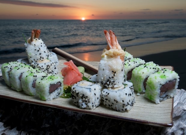 A platter of sushi is presented on a beach setting at sunset. The sushi rolls are topped with shrimp and garnished with pickled ginger, placed alongside a pair of chopsticks. The vibrant colors of the sky create a serene backdrop.