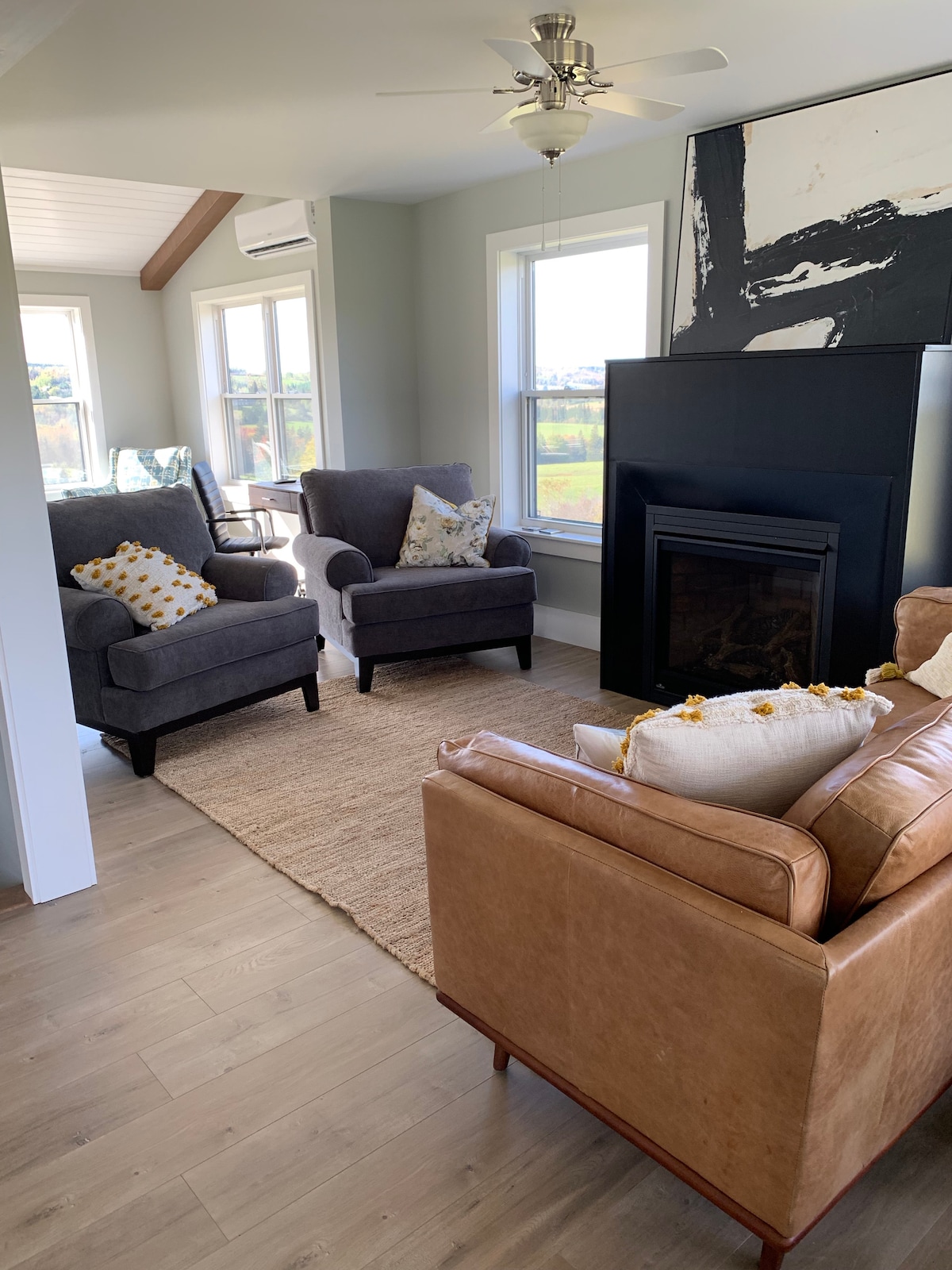 A cozy living area is featured with two grey armchairs adorned with patterned cushions. A leather sofa, positioned next to a sleek black fireplace, adds warmth. Natural light is provided by large windows, revealing views of the surrounding countryside, alongside a light-colored area rug on the floor.