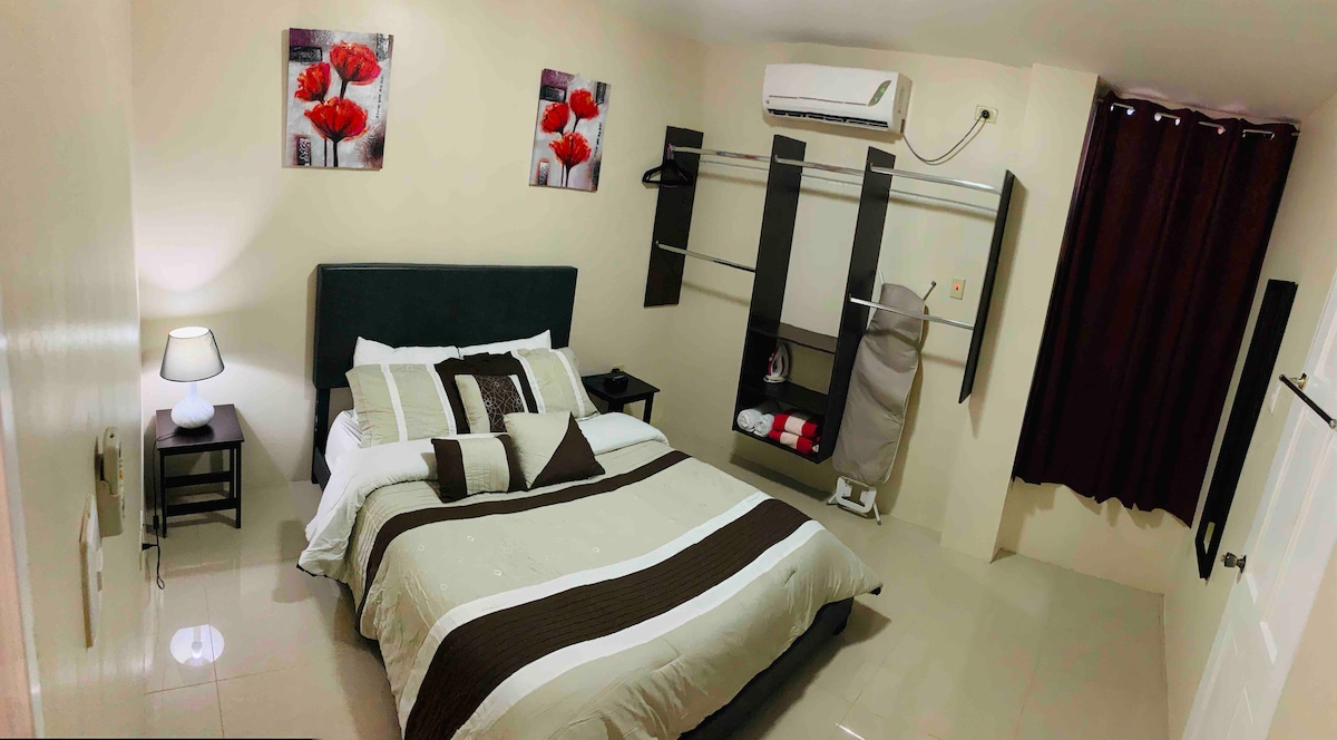 A well-appointed sleeping area features a spacious bed dressed in neutral-toned linens, with decorative artwork of red flowers above. A nightstand with a lamp is positioned beside the bed. A clothing rack and a soft armchair are included, while a dark curtain regulates light from the window.