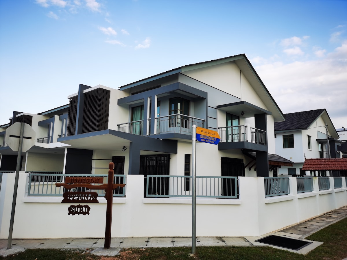 A modern two-story house features a combination of gray and white exterior walls. The entrance is marked by a wooden sign displaying the property name. A balcony extends from the upper floor, providing outdoor space. Surrounding sidewalks and a manicured lawn enhance the entrance.