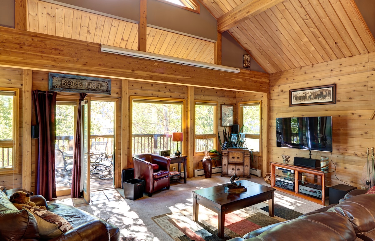 A spacious living area is enhanced by wooden beams and large windows allowing natural light. Comfortable seating is arranged around a central coffee table, with a television mounted on the wall. A door opens to a deck space with views of the surrounding nature.