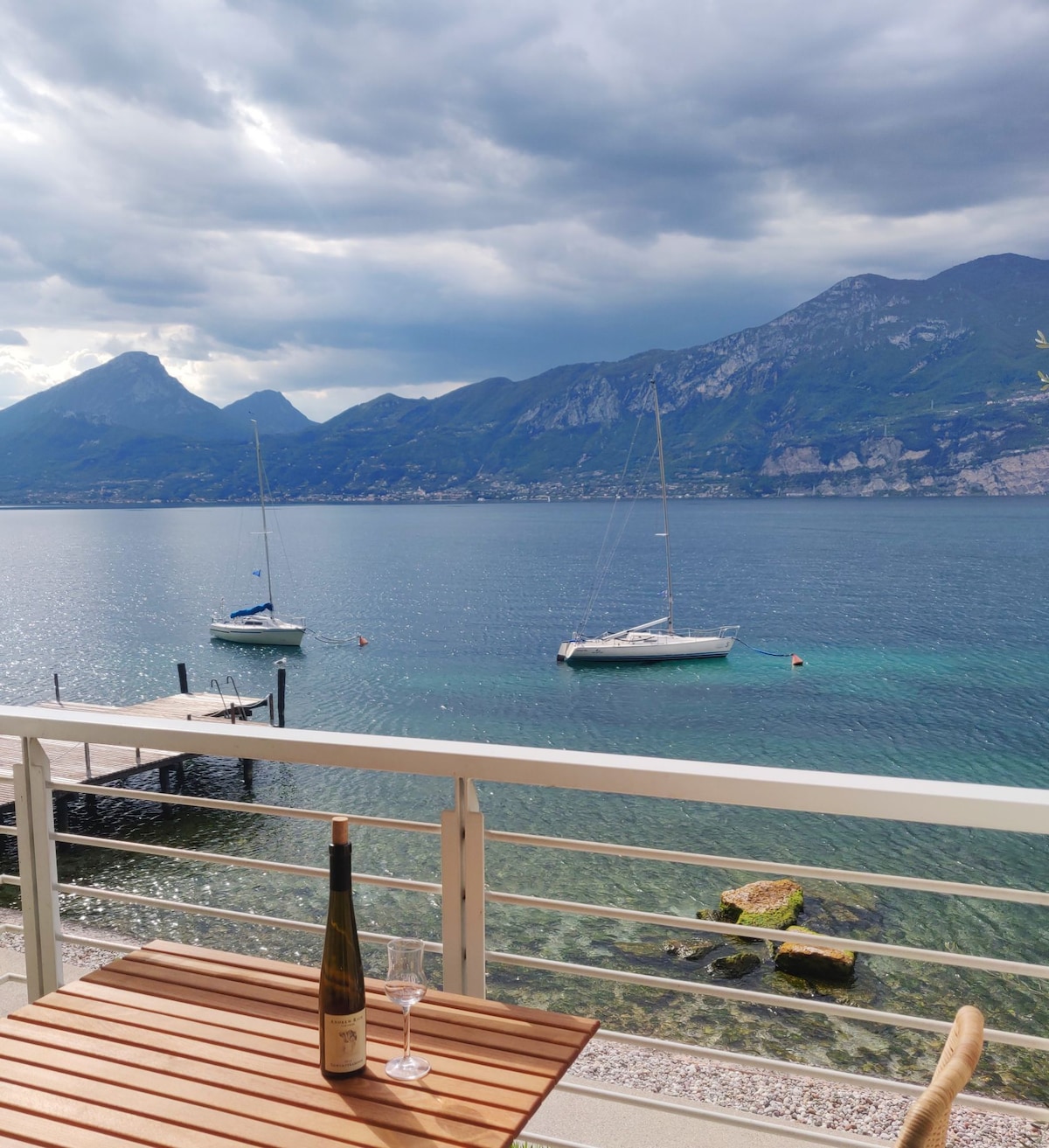 A private terrace offers a view of the lake, where two sailboats float on calm waters. The backdrop features distant mountains under a partly cloudy sky. A wooden table holds an open bottle of wine and a glass, inviting relaxation.
