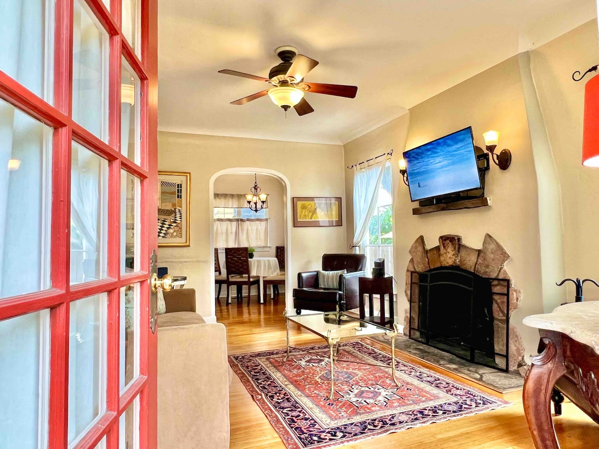 A welcoming living area is visible through red French doors. A ceiling fan hangs above, and a flat-screen television is mounted on the wall. A decorative rug lies on polished hardwood floors, complemented by a stone fireplace and comfortable seating arrangements to create an inviting space.