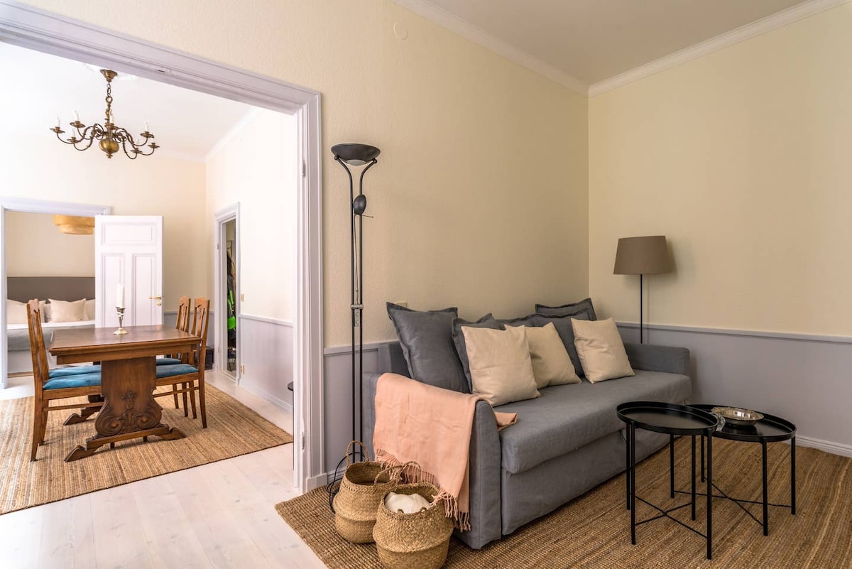 A living room area features a comfortable sofa adorned with throw pillows, complemented by a wooden coffee table. Light-colored walls and a jute rug add to the space's warmth, while an expansive dining area is visible through the doorway, showcasing a large wooden dining table and blue chairs.