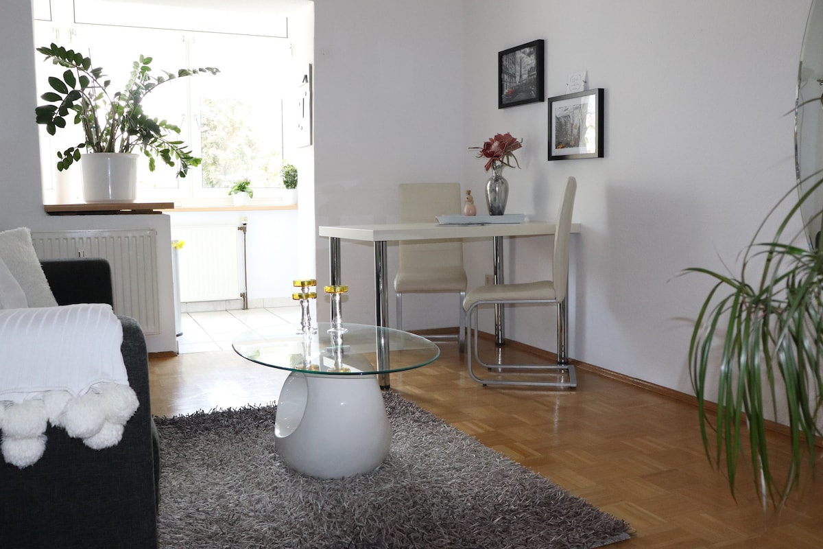 A well-lit dining area features a small glass table surrounded by two light-colored chairs. A decorative vase with flowers is placed on the table. A soft rug covers the wooden floor, and large windows allow natural light to brighten the space, accompanied by greenery from nearby plants.