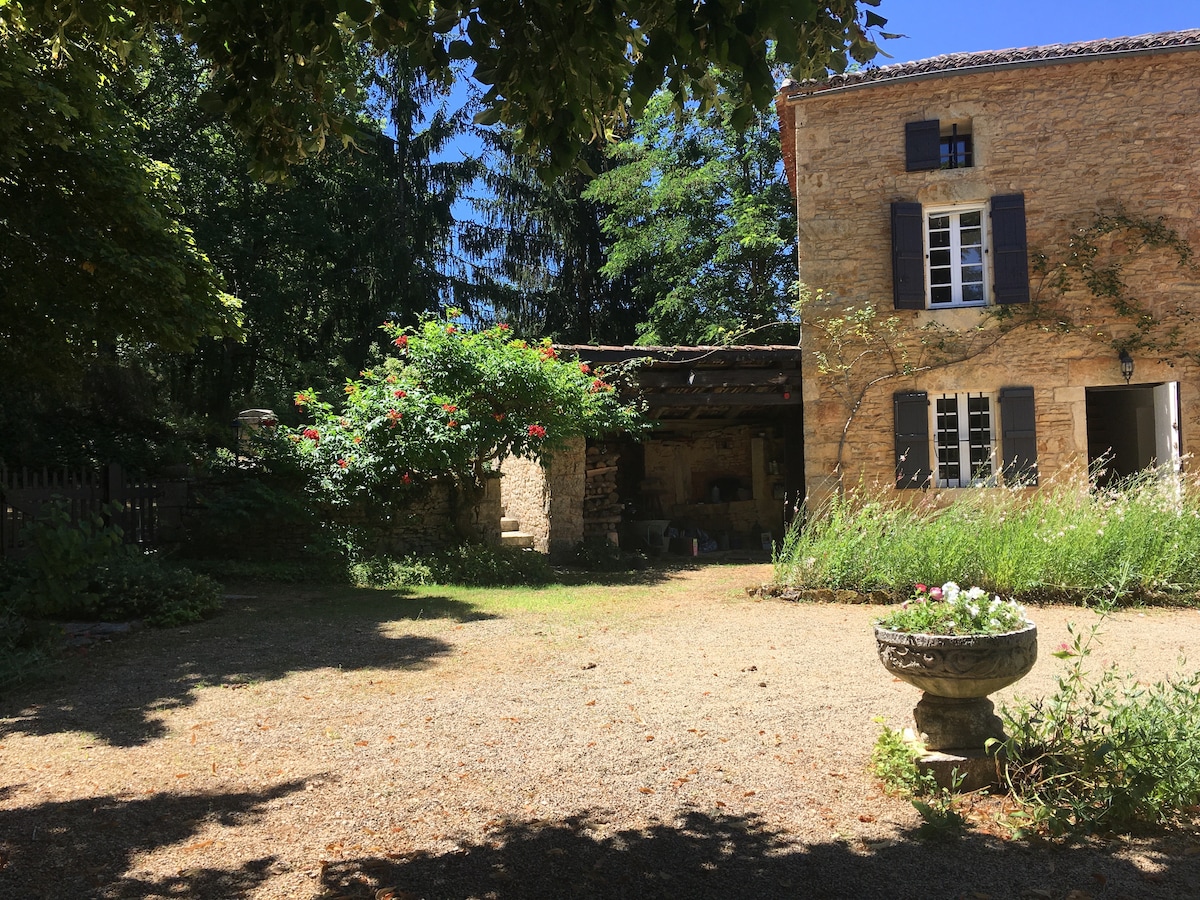 The exterior of a stone house is surrounded by lush greenery, with vibrant flowers near the entrance. A shaded area provides an inviting space along the house, marked by exposed stone walls and charming windows. A decorative stone planter is visible in the foreground.