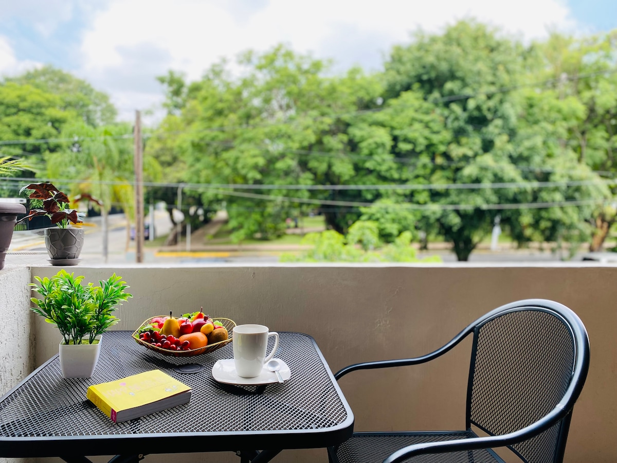 A small balcony features a round table set with a white plate of assorted fruits and a cup beside a yellow book. Lush greenery from nearby trees is visible in the background, providing a serene view of the surroundings.