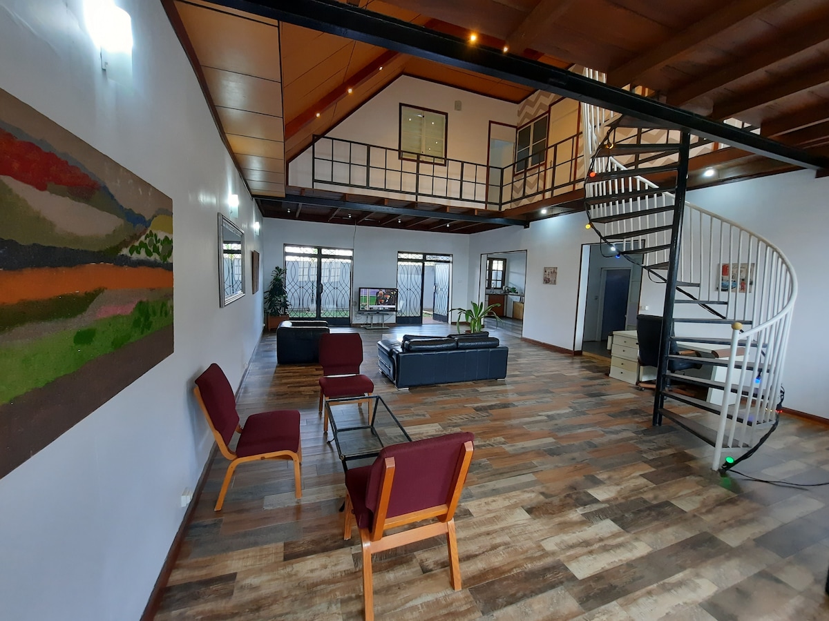 A spacious living area features a high ceiling with a lofted space above. Comfortable seating includes two burgundy armchairs and a sectional sofa, arranged around a coffee table. A spiral staircase leads to the upper level. Natural light enters through multiple large windows, enhancing the open design.