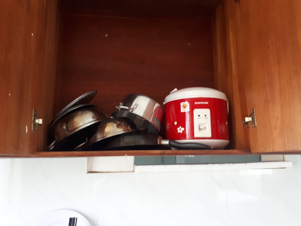 A wooden cabinet is displayed with several stacked metal pots and a red rice cooker. The interior appears organized, with the pots showing varied sizes and the rice cooker having a clear front panel. The cabinet's clean design complements the kitchen's overall functionality.