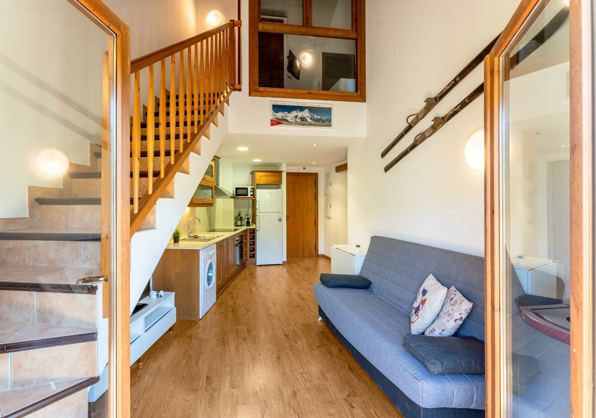 An open living space features a gray sofa positioned by a staircase leading to an upper level. A compact kitchen area is visible with wooden cabinetry, appliances, and a dining space. Light wood flooring enhances the airy feel of the room.