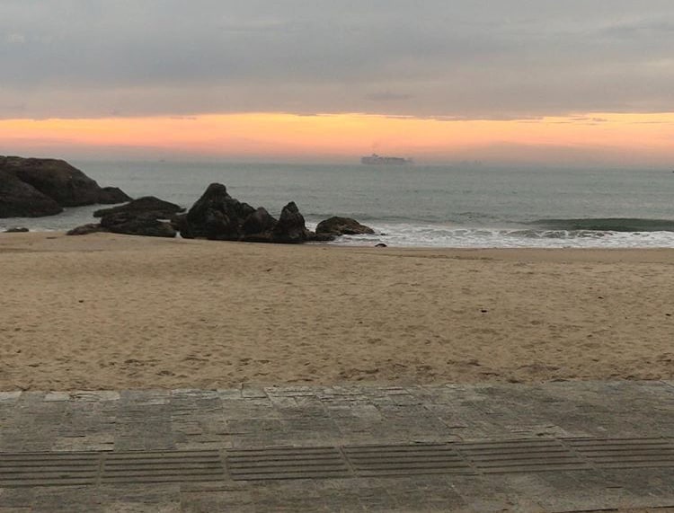 A sandy beach is viewed, stretching along the shoreline. Gentle waves lap at the shore, with scattered rocks in the foreground. A calm sea meets a pastel-colored sky at sunset, creating a serene atmosphere. The image captures the natural beauty of the coastal landscape.