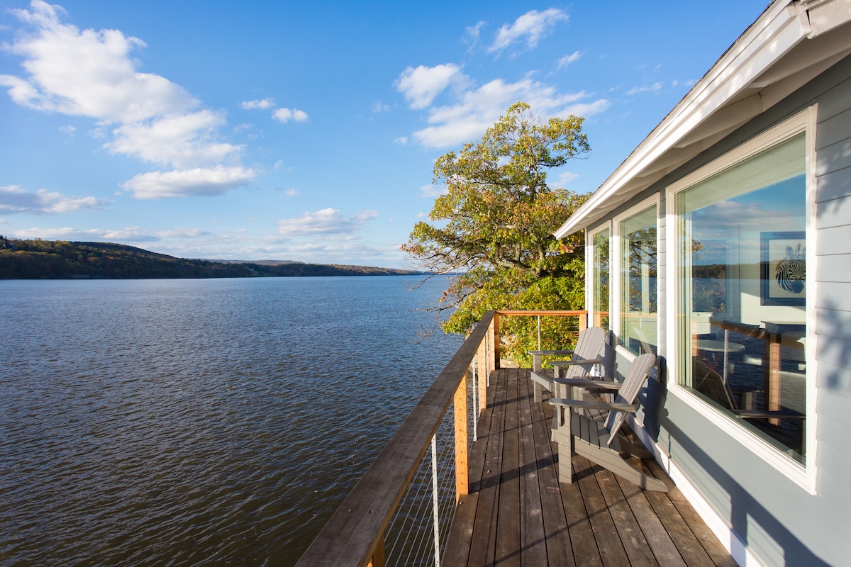A wraparound deck is shown overlooking the calm river, with two adirondack chairs positioned for views. The expansive glass windows of the cottage reflect the serene water and scenic landscape, under a blue sky adorned with scattered clouds.