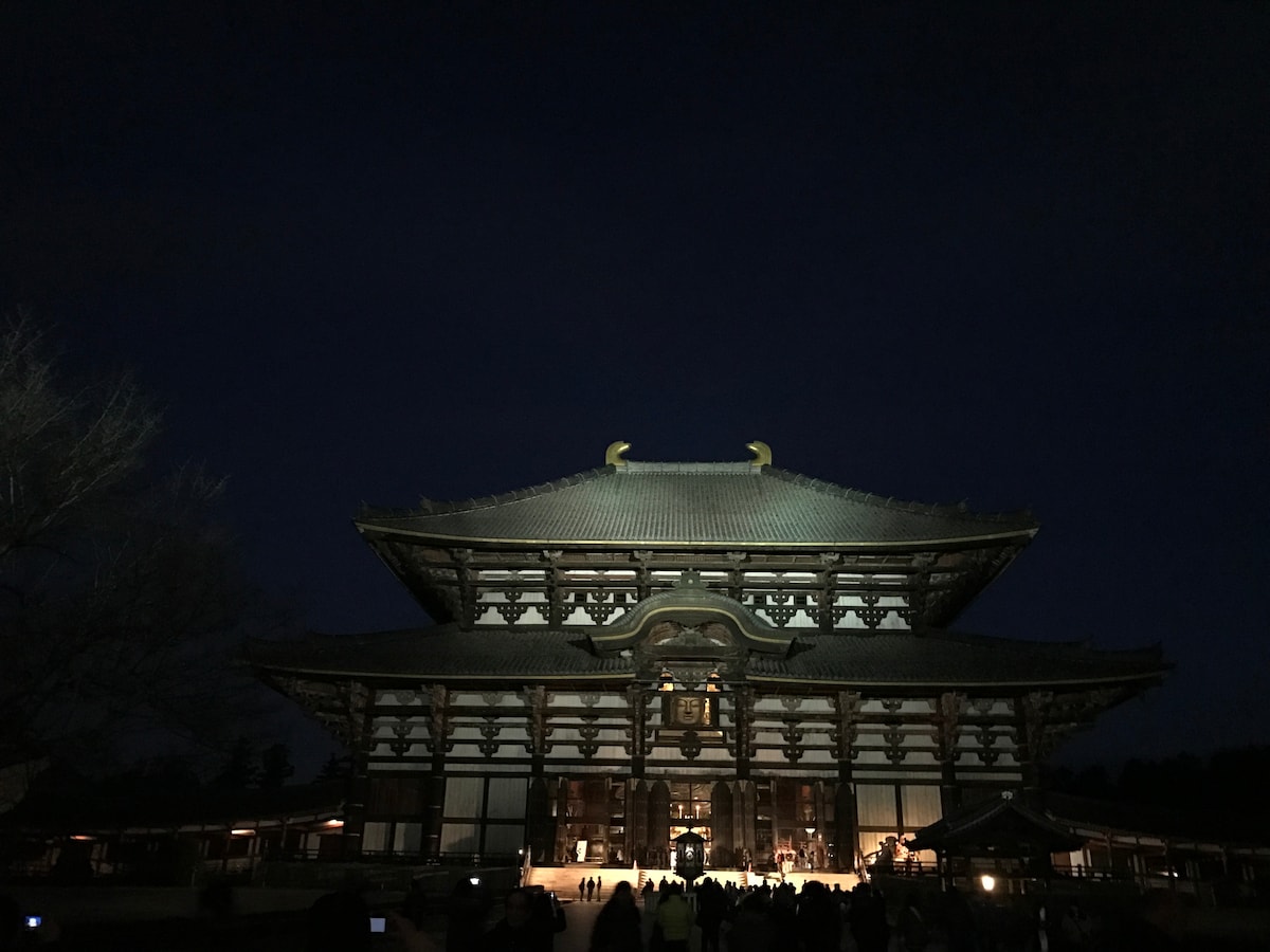The illuminated Todaiji temple stands grandly against the night sky, showcasing intricate architectural details. The warm light casts a glow on the entrance and surrounding structures, highlighting the peaceful atmosphere created by visitors gathered in the foreground.
