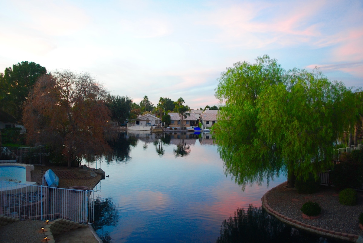 A serene lake view is captured, showcasing calm waters reflecting the pastel sky. Lush trees line the banks, with a nearby section featuring landscaped rocks. Residential homes are visible in the background, enhancing the peaceful ambiance of the setting.