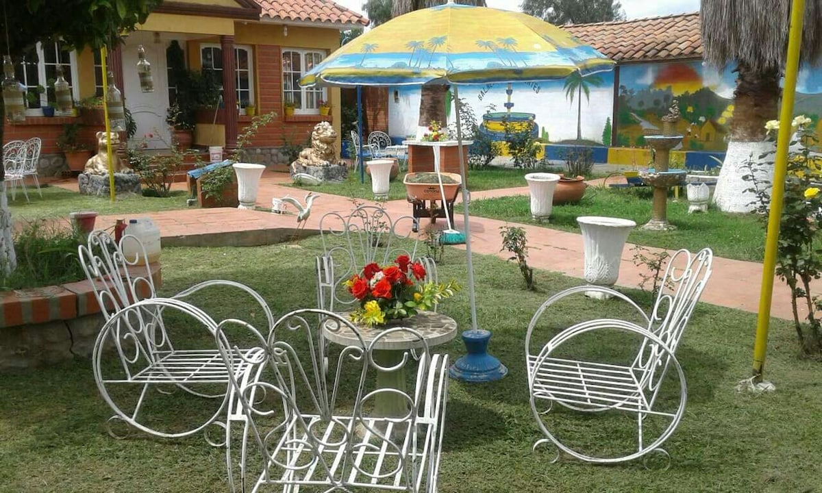 A charming outdoor area features several white, wrought-iron chairs arranged around a small table adorned with a vibrant floral centerpiece. The space is surrounded by well-maintained grass and colorful landscaping, with decorative pottery and a mural visible in the background.