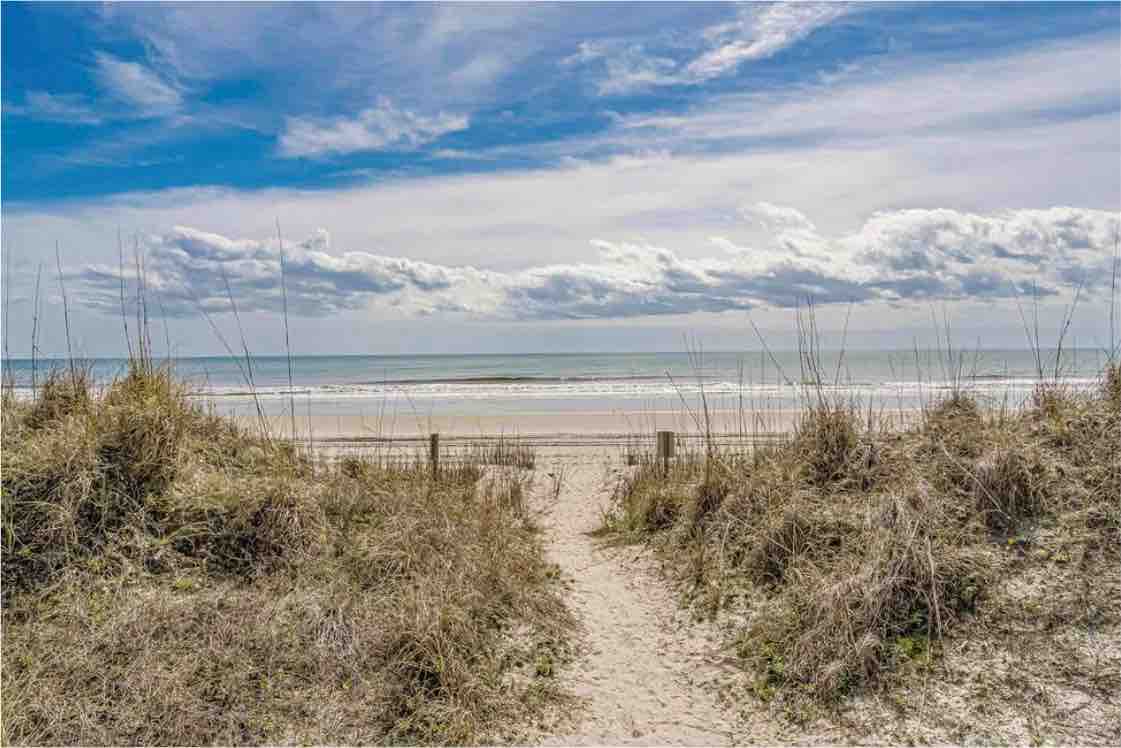A sandy path leads through beach grass toward the shoreline, where the ocean meets the sky. Gentle waves can be seen lapping at the beach, under a partly cloudy sky that adds a serene backdrop to the coastal scene.