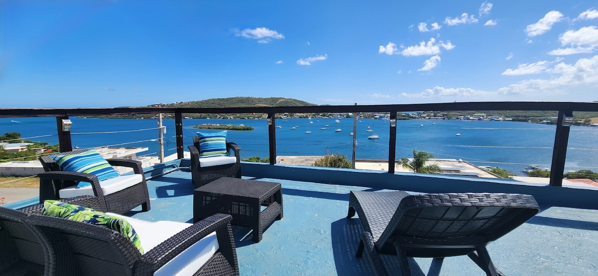 An outdoor terrace offers comfortable seating with stylish chairs and a coffee table, overlooking Ensenada Honda Bay. The clear blue sky and water create a serene atmosphere, while boats are visible in the bay, enhancing the view.