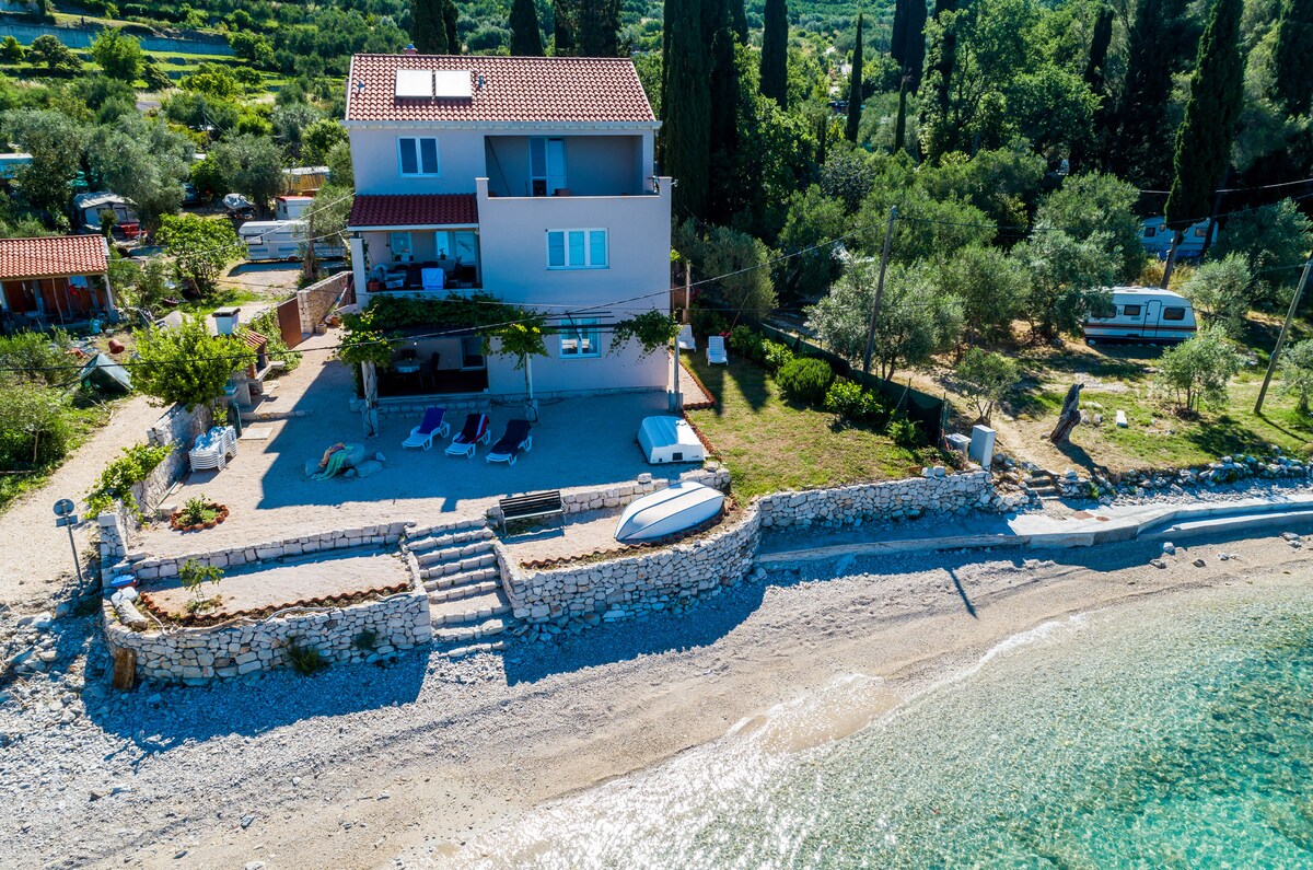 The accommodation is situated directly on the beach, featuring a sandy shore that transitions to clear water. An outdoor seating area with loungers is visible, and olive trees surround the property, providing shade. The two-story building showcases a balcony with partial sea views.