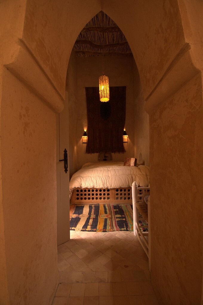A softly lit entrance leads into a cozy room featuring a spacious bed adorned with neutral bedding. The room displays traditional decor, including a woven wall hanging and ambient lighting, while the arched doorway emphasizes the room's inviting atmosphere.