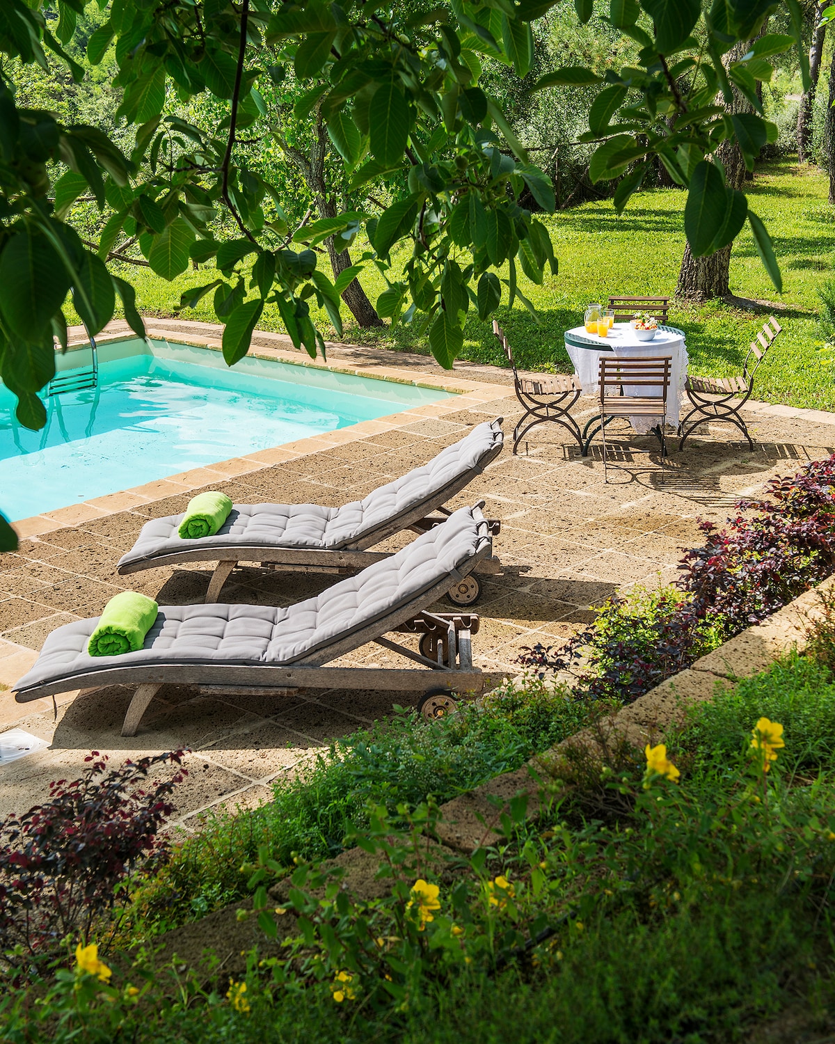 A serene outdoor space features a turquoise pool surrounded by lush greenery. Two comfortable lounge chairs with soft cushions are positioned nearby, while a dining table set for four with bright tableware invites relaxation. Colorful flowers enhance the vibrant garden atmosphere.