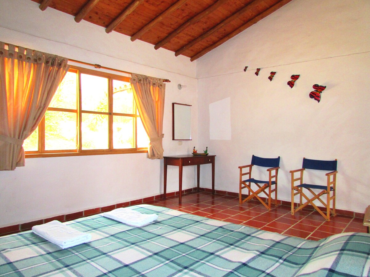 A spacious room features two chairs positioned near a large window, allowing natural light to fill the space. A wooden table is present opposite the window, and fresh linens are neatly arranged on the floor. A charming decorative piece hangs on the wall.